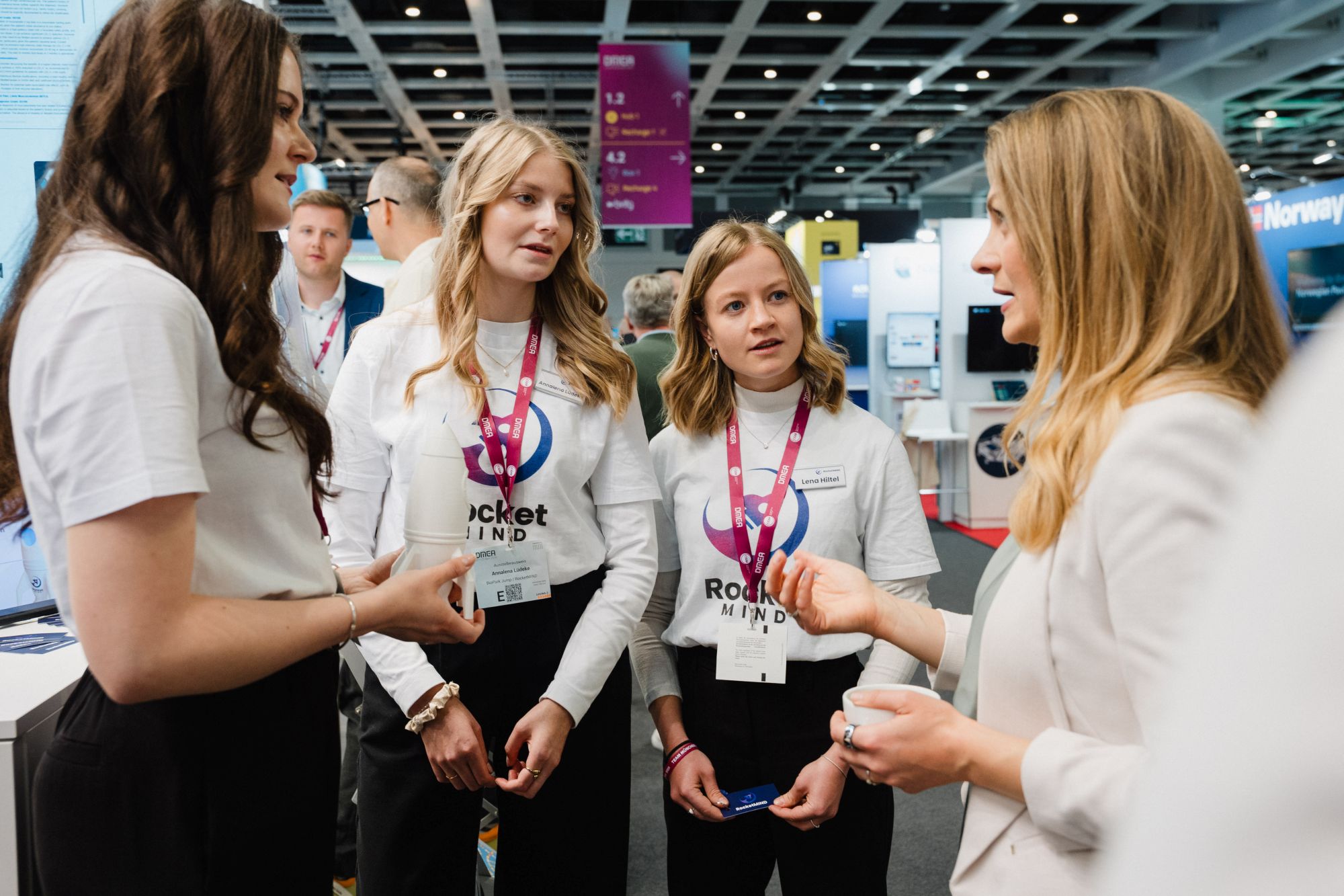 Vier Frauen im Gespräch auf einer Messe, drei davon tragen weiße T-Shirts mit dem Schriftzug "Rocket MIND" und Namensschilder an roten Bändern