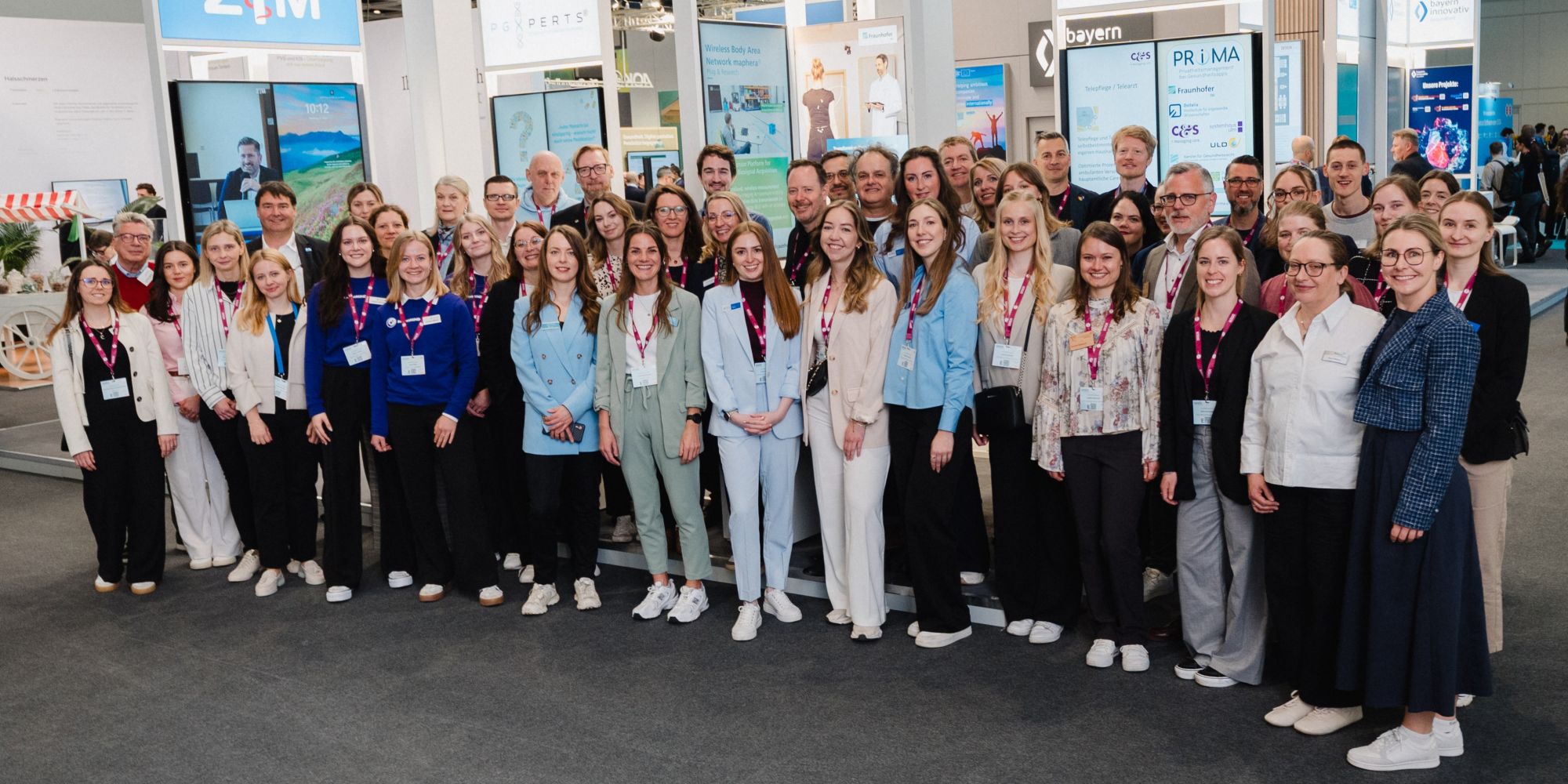 Große Gruppe von Menschen steht in einer Messehalle vor Messeständen und lächelt in die Kamera