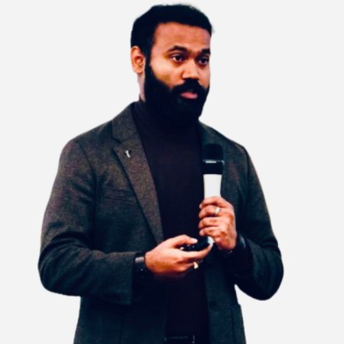 Mann mit Schwarzen Haaren und schwarzem Vollbart in dunkelgrauem Sakko und Schwarzem Oberteil