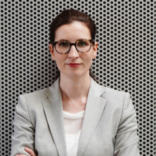 Frau mit braunen Haaren und Zopf, Brauer Brille und weißem Tshirt mit grünem Blazer vor einer Wand mit vielen Löchern