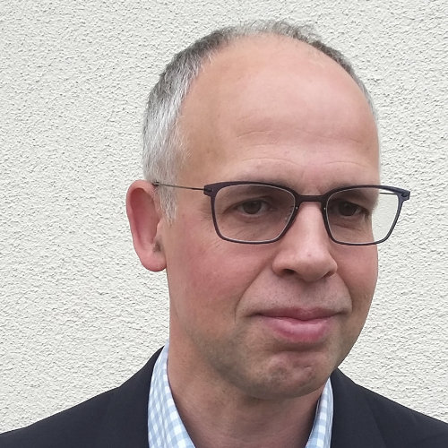 Das Bild zeigt einen Mann mit einer schwarzen Brille in Hemd und Sakko