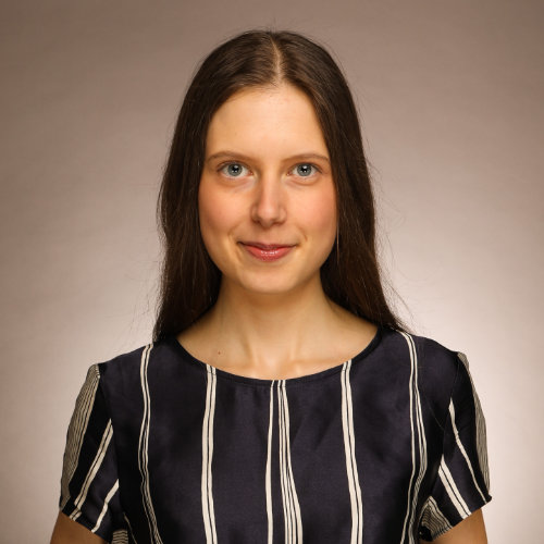 Portraitbild von Junianna Zatsarnaja, einer Frau mit braunen langen Haaren und einem schwarzen Oberteil mit weißen streifen vor einem hellbraunen Hintergrund