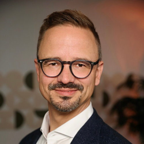 Das Bild zeigt einen Mann mit schwarzer Brille und einem Bart, der ein weißes Hemd und ein schwarzes Sakko trägt