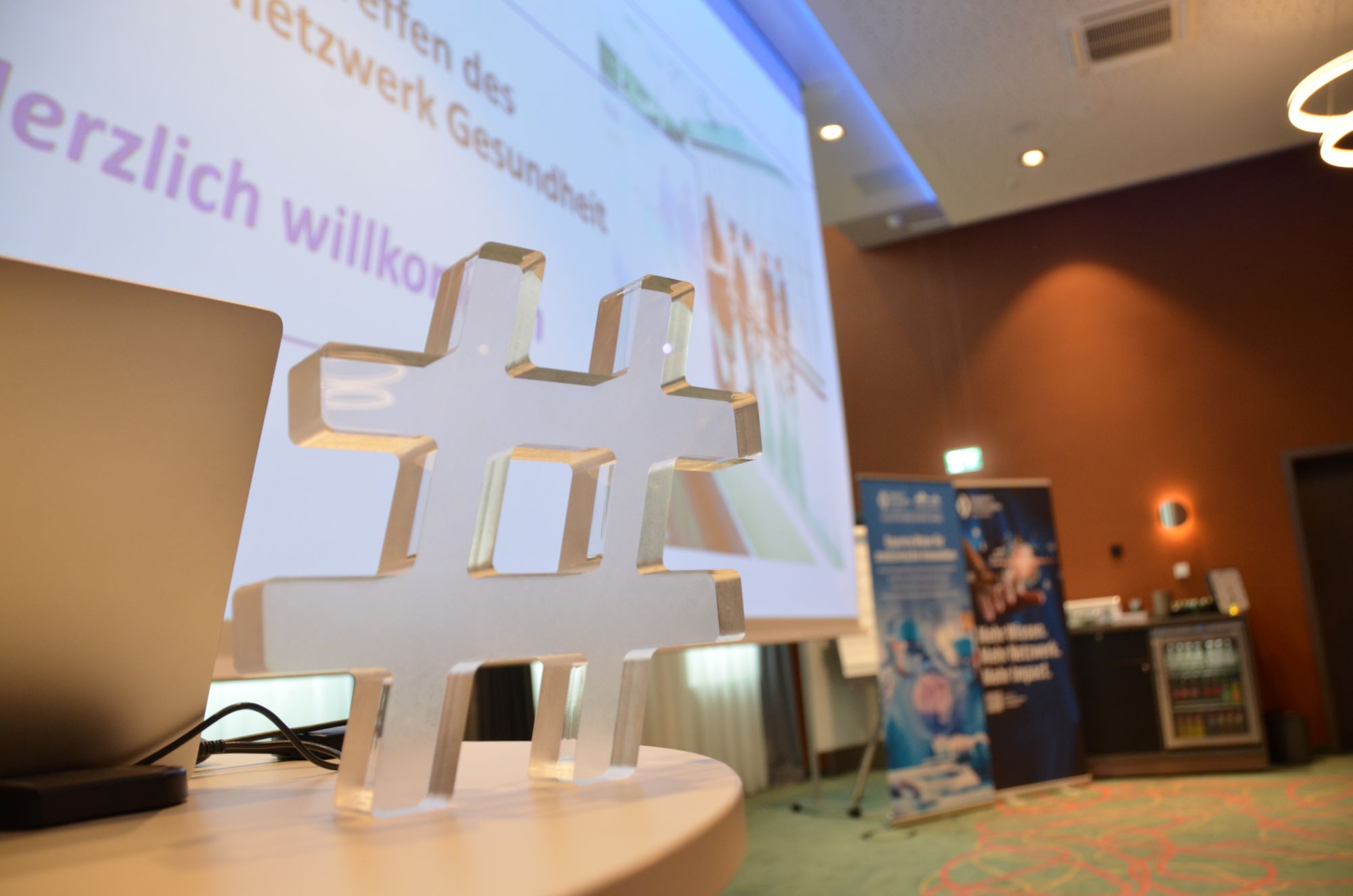 Großes weißes Hashtag-Symbol auf einem Tisch vor einer Projektionswand in einem Konferenzraum