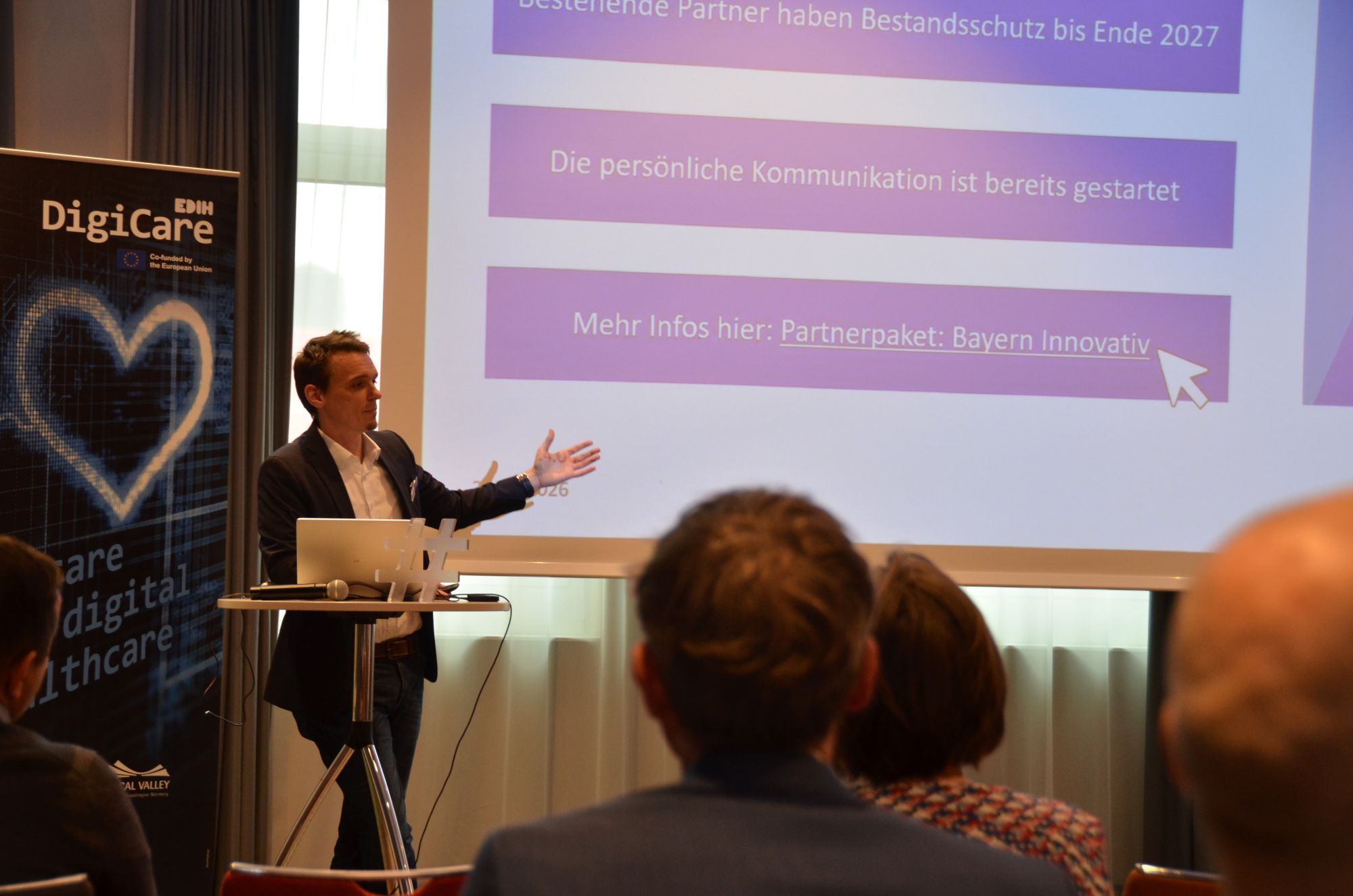Person steht vor Leinwand mit Präsentation und zeigt auf Textabschnitt, Publikum sitzt im Vordergrund