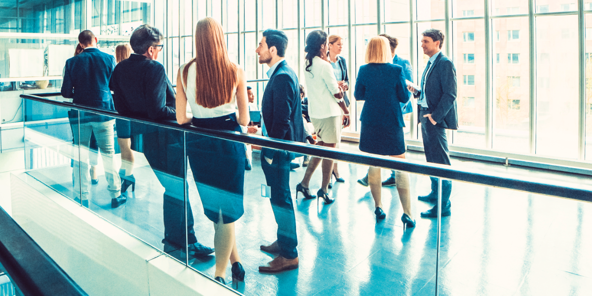 Das Bild zeigt eine Gruppe von Geschäftsleuten, die sich in einem modernen Bürogebäude aufhalten. Sie stehen an einem Geländer, das auf einen unteren Bereich des Gebäudes blickt. Einige der Personen unterhalten sich, andere schauen nachdenklich aus dem Fenster