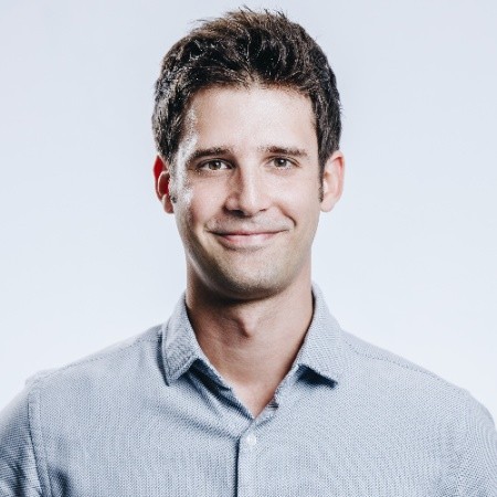 Junger Mann mit kurzem, dunklem Haar trägt ein hellblaues Hemd mit offenem Kragen vor hellem Hintergrund