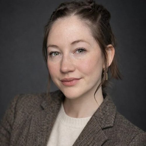 Person mit zusammengebundenem Haar trägt braunen Blazer und hellen Pullover vor dunklem Hintergrund