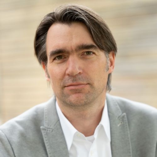 Mann mit grauem Sakko und weißem Hemd vor unscharfem Hintergrund
