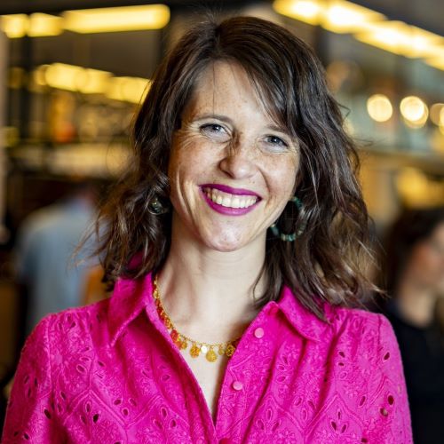 Frau mit schulterlangem gewelltem Haar trägt eine pinke Bluse mit Lochmuster und eine goldene Halskette mit runden Anhängern in einem hell erleuchteten Innenraum