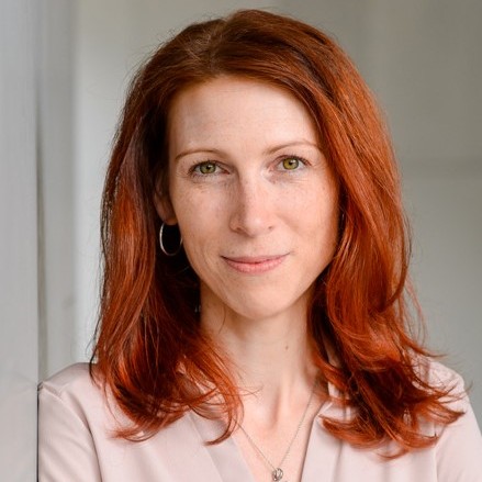 Frau mit schulterlangem, gewelltem roten Haar trägt silberne Creolen und eine Kette mit kleinem Anhänger vor hellem Hintergrund