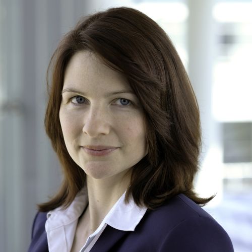 Frau mit schulterlangem braunem Haar trägt ein weißes Hemd und ein dunkles Sakko vor einem unscharfen Fensterhintergrund