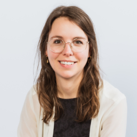 Frau mit schulterlangem braunem Haar trägt schwarzen Pullover und helle Strickjacke vor hellem Hintergrund