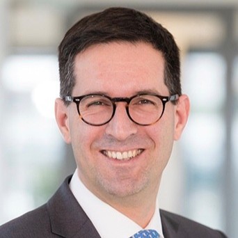 Mann mit dunklem Haar trägt dunklen Anzug, hellblaues Hemd und hellblaue Krawatte vor unscharfem Hintergrund