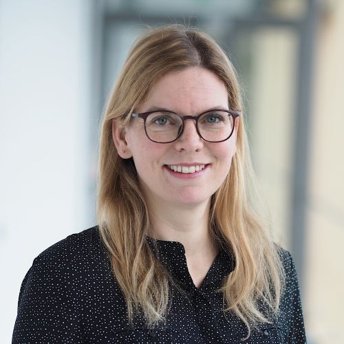 Frau mit langen blonden Haaren trägt eine schwarze Bluse mit weißem Punktmuster vor unscharfem Hintergrund