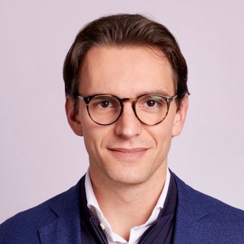 Oberkörperporträt eines Mannes mit braunen Haaren, der einen weißen Hemdkragen, einen dunklen Pullover und ein blaues Sakko trägt