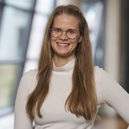 lächelnde junge Frau mit langen Haaren und Brille vor einer modernen Glasfasade 