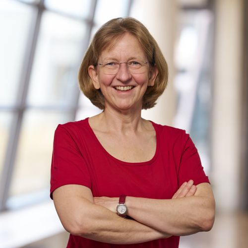 lächelnde Frau mit mittelblonden Haaren und Brille vor einer modernen Glasfasade