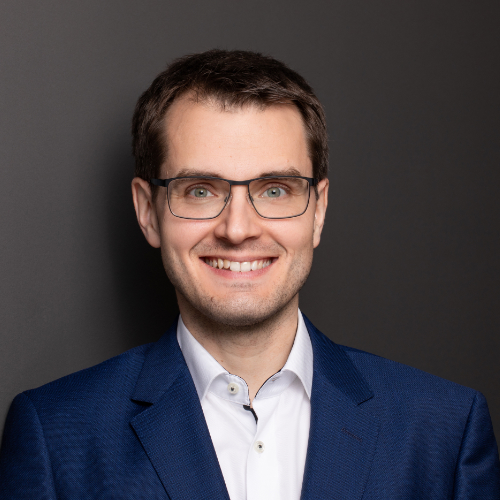 Ein Mann mit Brille und blauem Anzug lächelt freundlich in die Kamera.