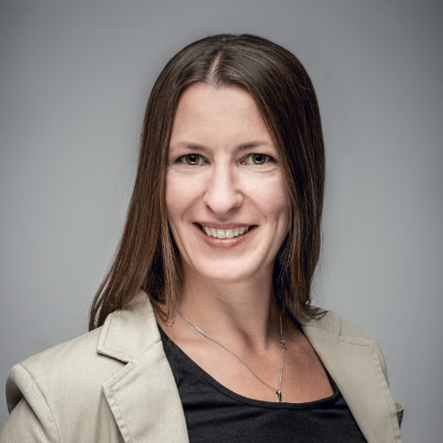Das Bild zeigt eine dunkelhaarige Frau, die ein dunkles Oberteil und einen beigen Blazer trägt