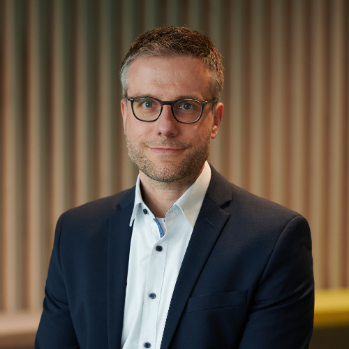 Das Bild zeigt einen Mann mit schwarzer Brille, der ein weißes Hemd und ein dunkelblaues Sakko trägt