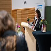 Frau steht am Rednerpult mit Mikrofon und hält Notizen, im Hintergrund Banner mit Schriftzug 'Zukunft Pflege', im Vordergrund unscharfe Person und Blumenarrangements auf Tischen