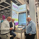 Auf dem Bild sind drei Herren zu sehen, welche vor einem Messestand posieren.