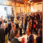 "Viele Personen befinden sich in einem gut beleuchteten Veranstaltungsraum. Die Anwesenden stehen in Gruppen zusammen und unterhalten sich. Einige Tische sind sichtbar. Die Personen tragen formelle oder geschäftliche Kleidung."