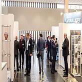 Das Bild zeigt eine Gruppe von Personen in einem Flur oder Ausstellungsbereich, vermutlich im Rahmen einer Konferenz oder Messe. Auf beiden Seiten befinden sich Stände oder Displays, und die Personen wirken in Gespräche oder die Betrachtung der Ausstellungen vertieft. 