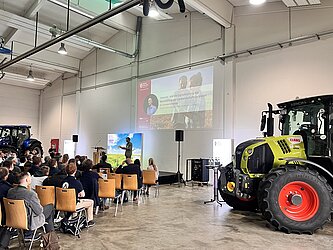 Teilnehmende der Veranstaltung Landwirtschaft im Wandel die in den Vortragsreihen sitzen und einem Vortrag zuhören. Daneben steht ein Traktor als Ausstellungsstück