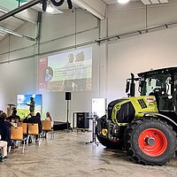 Teilnehmende der Veranstaltung Landwirtschaft im Wandel die in den Vortragsreihen sitzen und einem Vortrag zuhören. Daneben steht ein Traktor als Ausstellungsstück