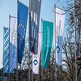 Das Bild zeigt eine Reihe von Fahnen an Fahnenmasten. Zu sehen sind unter anderem die Flagge der Europäischen Union, eine blau-weiß karierte Fahne, eine weiße Fahne mit blauer Schrift sowie weitere Fahnen mit unterschiedlichen Designs und Farben.