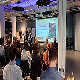 Auf diesem Bild ist eine Gruppe von Menschen zu sehen, die in einem Konferenz- oder Seminarraum sitzen und auf einen Präsentationsbildschirm schauen. Vorne im Raum stehen Präsentatoren, und auf dem Bildschirm werden Folien mit Text und Grafiken angezeigt. 