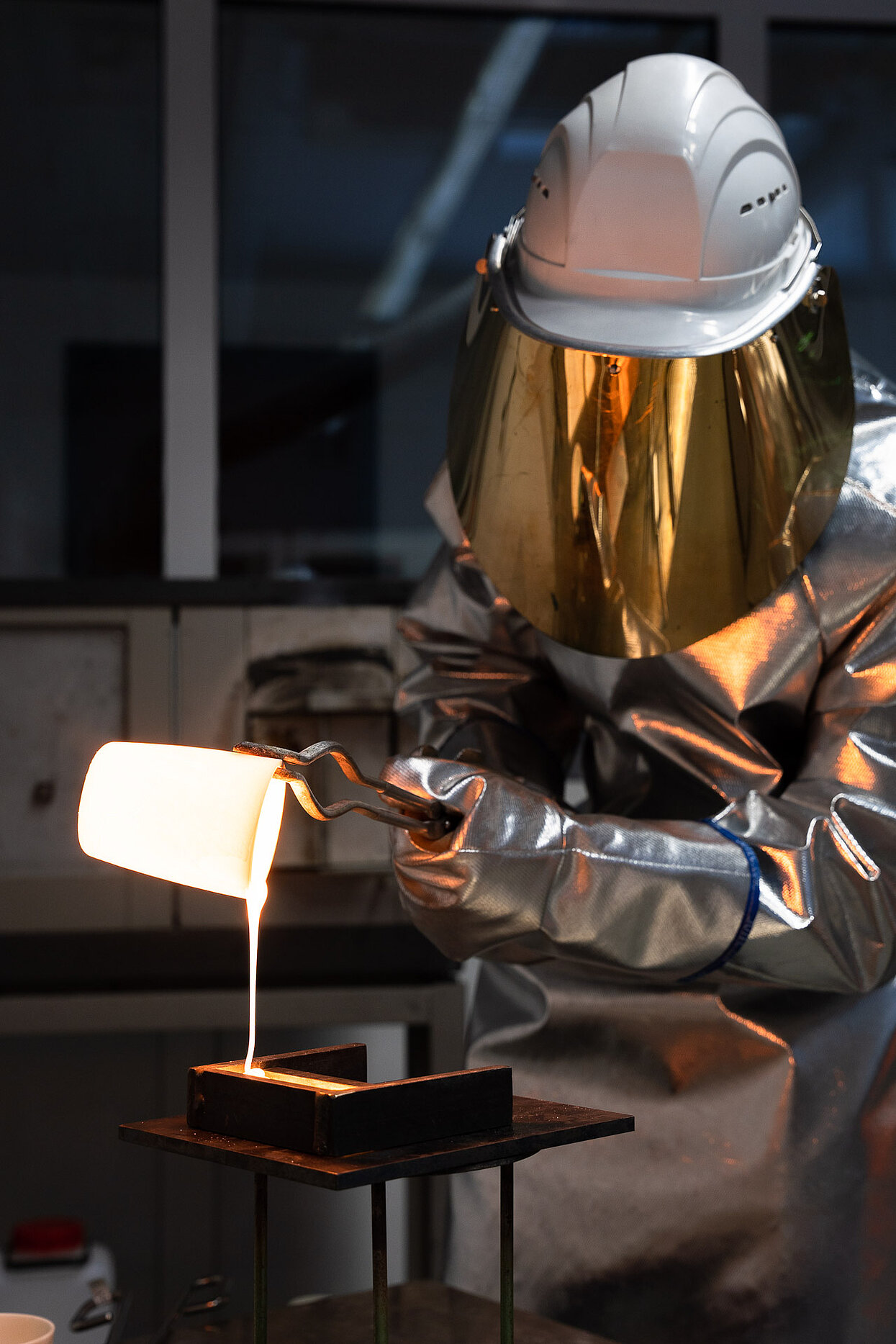 Mitarbeiter in Schutzanzug und Helm gießt glühend flüssiges Metall aus einem Tiegel in eine Form.