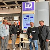 Auf dem Bild sind vier Personen zu sehen, welche vor einem Messestand in lila Farbe posieren.