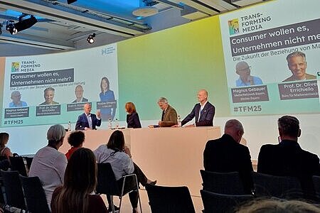 Auf der Bühne ist ein Pult aufgebaut, hinter dem vier Personen stehen. Sie führen eine Paneldiskussion über die Zukunft der Beziehung zwischen Marke und Haltung. Die Namen der Personen sind im Hintergrund eingeblendet. Vom Publikum sind lediglich die Umrisse erkennbar. 