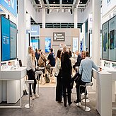 Mehrere Personen sitzen und stehen an hohen Tischen in einem hellen Messestand mit Bildschirmen und dem Schriftzug 'bayern innovativ' im Hintergrund