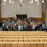 Große Gruppe von Menschen steht in einem Hörsaal vor leeren Sitzreihen und blickt zur Kamera