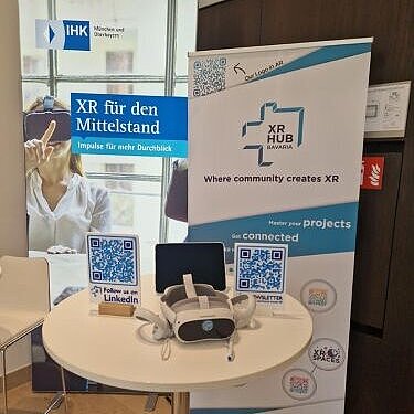 Auf einem runden Tisch liegen eine VR-Brille und mehrere QR-Codes, dahinter ste-hen zwei Roll-up-Banner mit Informationen zu XR und dem XR Hub Bavaria.