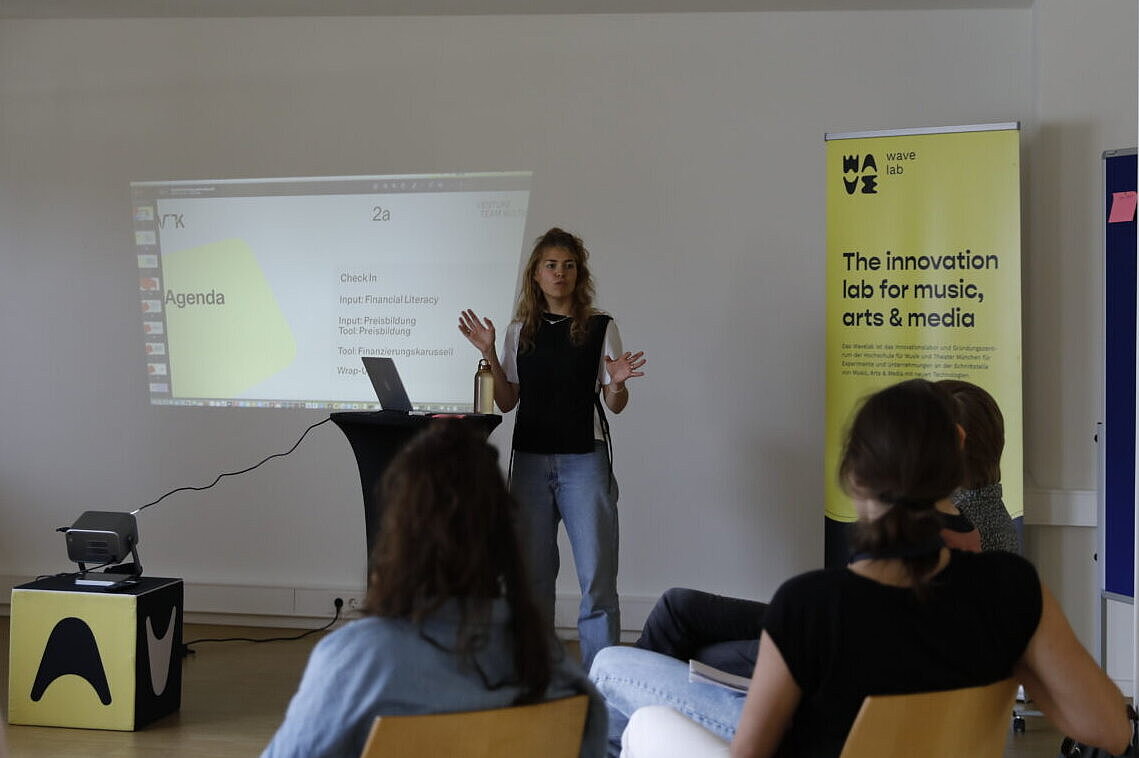 Zu sehen ist eine weibliche Person, wie sie auf der Bühne stehend einen Vortrag hält. Eine Folie ist durch einen Beamer sichtbar. Rechts neben ihr steht ein gelbes Roll-Up mit der Aufschrift "The Innovation lab for music, arts & media". 