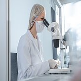 Person mit Haarnetz und Schutzbrille sitzt an Mikroskop in Laborumgebung
