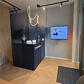 Empfangsbereich mit dunklem Tresen, moderner Beleuchtung und Bildschirm an der Wand.