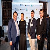 Vier Personen stehen vor einem Hintergrund mit verschiedenen Logos und Schriftzügen. 