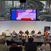 Panel: Mathis Nitschke, Komponist, Technologe und Unternehmer bei der Sofilab GmbH in München, TJ Gräbe, Problemlöser bei der isento GmbH, Prof. Daniel Nusser, Professor und Studiengangsleitung für Prozess- und Produktdesign an der DIPLOMA Hochschule, sowie Dr. Jürgen Baier, Geschäftsführer der isento GmbH, sitzen vorne in Sesseln nebeneinander und diskutieren vor Publikum. Moderiert wird die Runde von Johanna Lison, Projektmanagerin CreativeTechHUB, Bayern Innovativ GmbH.