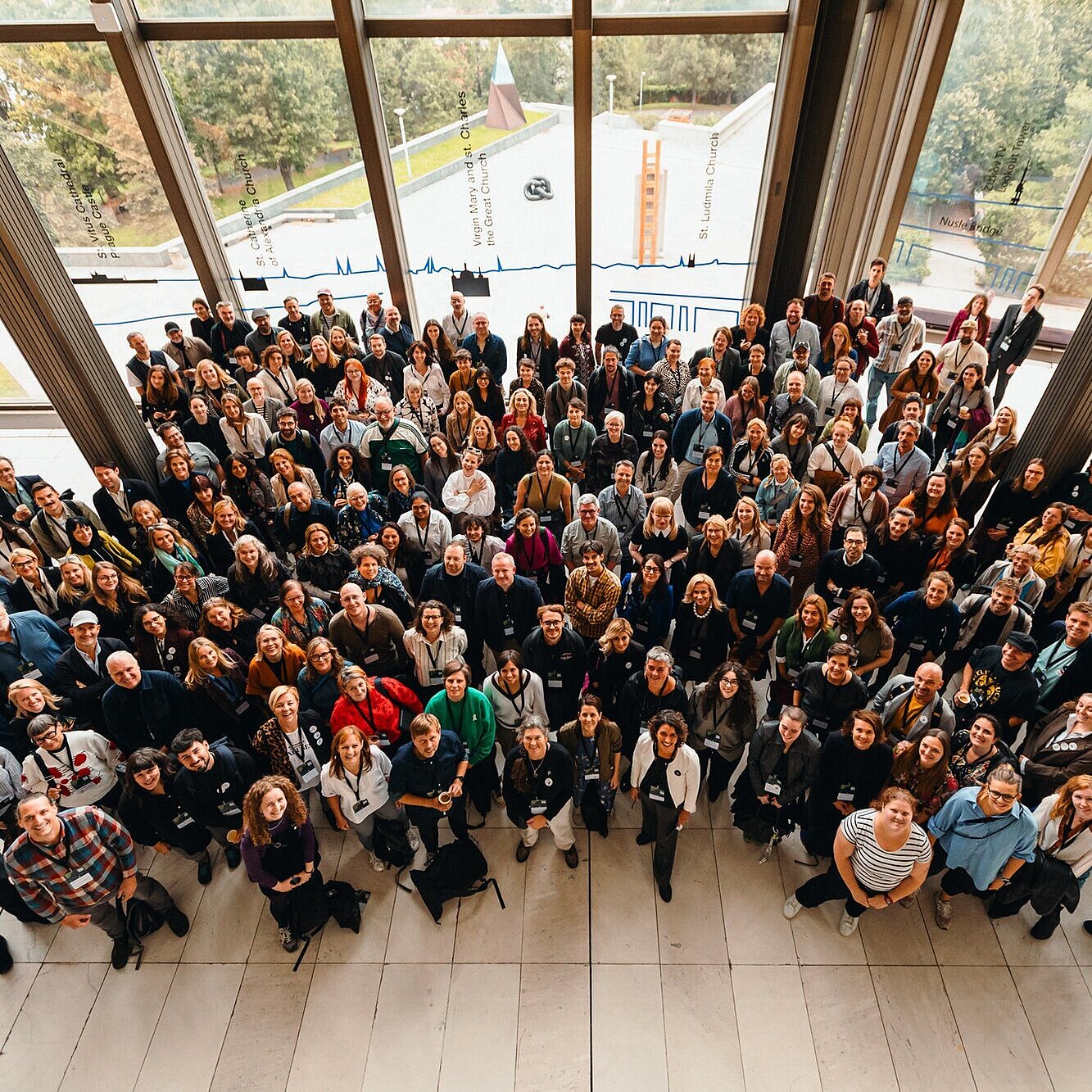 Ein von oben aufgenommenes Gruppenbild zeigt einige Menschen, die zur Kamera hinaufblicken. Sie sind in mehreren Reihen dicht beieinander angeordnet und lächeln. Durch die großen Fenster im Hintergrund sind Bäume und ein Teil eines Daches zu sehen. 