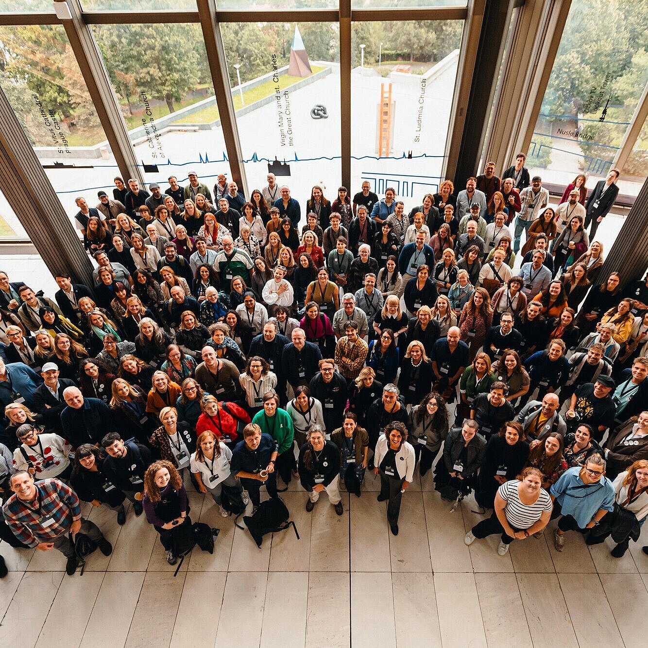 Ein von oben aufgenommenes Gruppenbild zeigt einige Menschen, die zur Kamera hinaufblicken. Sie sind in mehreren Reihen dicht beieinander angeordnet und lächeln. Durch die großen Fenster im Hintergrund sind Bäume und ein Teil eines Daches zu sehen. 