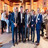 "Eine Gruppe von Personen in formeller Kleidung steht zusammen bei einer Veranstaltung in Innenräumen. Im Hintergrund sind weitere Gäste, einige Tische und Dekorationen bei warmer Beleuchtung zu sehen."