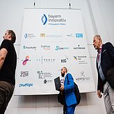 Großes Banner mit Logos verschiedener Unternehmen und Institutionen, darunter bayern innovativ, Fraunhofer, Hochschule Hof, gehalten von drei Personen in einem hellen Raum
