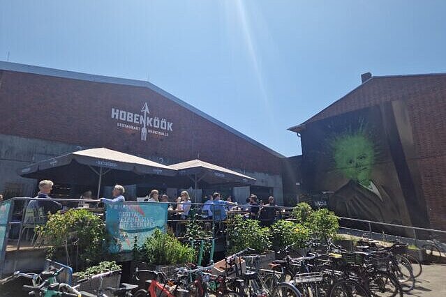 Zu sehen ist der Außenbereich der Veranstaltungslocation. Der Himmel is strahlend blau. Außerdem sind sitzende Menschen unter Sonnenschirmen zu sehen. Es wirkt nach einer geselligen Mittagspausen-Situation. 