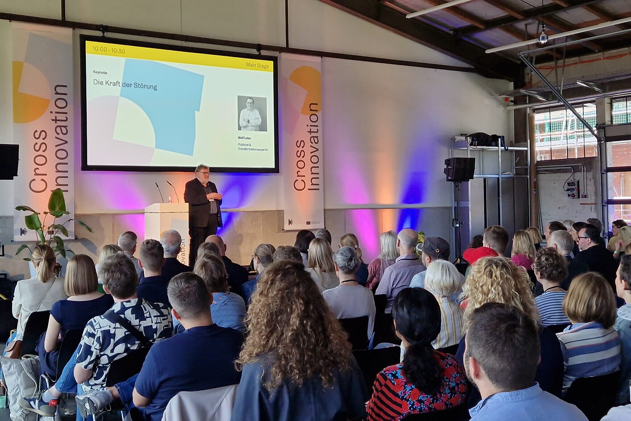 Zu sehen ist der Vortragsraum, während auf der Bühne ein Vortrag gehalten wird und die ZuhörerInnen gespannt hinhören. Details sind nicht zu erkennen, dafür wirkt die Atmosphäre durch warmes Licht sehr angenehm.